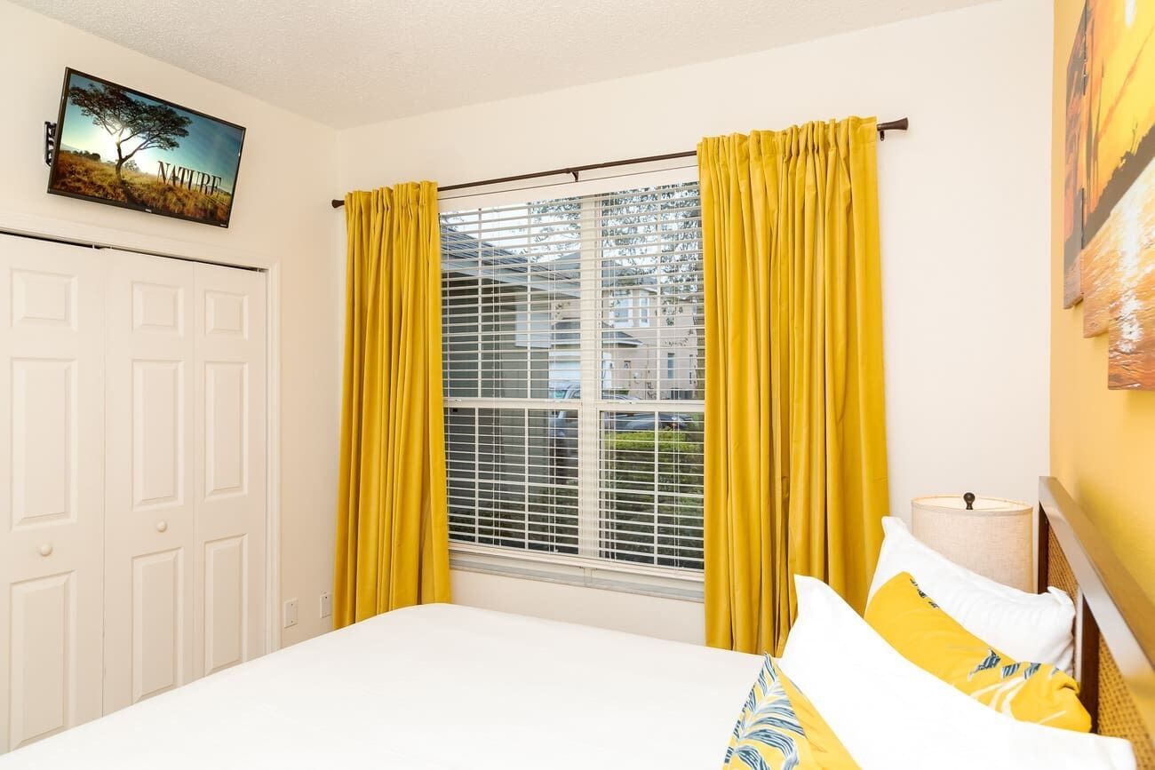 A bedroom with a bed, yellow curtains, a window with blinds, a painting on the wall, and a lamp on the bedside table.