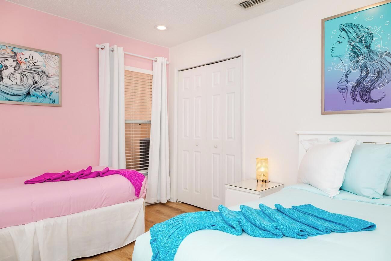 Un dormitori amb dos llits, un amb llençol rosa i l'altre amb llençol blau. Hi ha una porta blanca i una finestra amb cortines blanques.