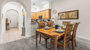 A dining area with a wooden table and chairs, a kitchen in the background, and a framed artwork on the wall. - Slice Of Sunshine At Caribe Cove (Kissimmee)