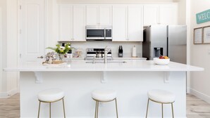 Una cocina moderna con muebles blancos, una isla central con taburetes, microondas, refrigerador y cafetera.