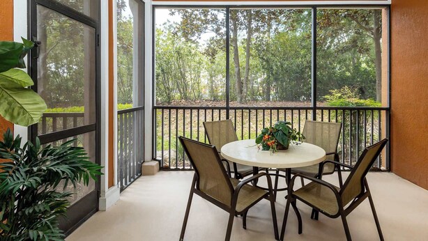 A covered patio with a round table and four chairs, surrounded by a railing, overlooking a garden with trees and shrubs.