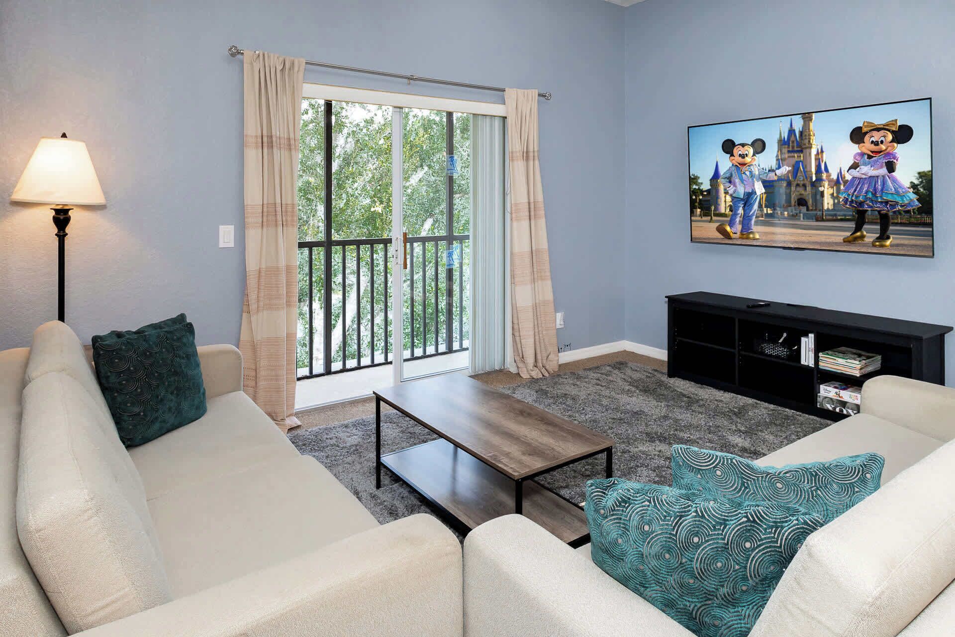 A living room with a sofa, a coffee table, a lamp, a TV, and a painting of Disney characters.