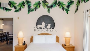 Habitación de hotel con cama, dos lámparas de noche, un cuadro en la pared con un león y un cerdo, y una decoración de hoja tropical sobre el cuadro.