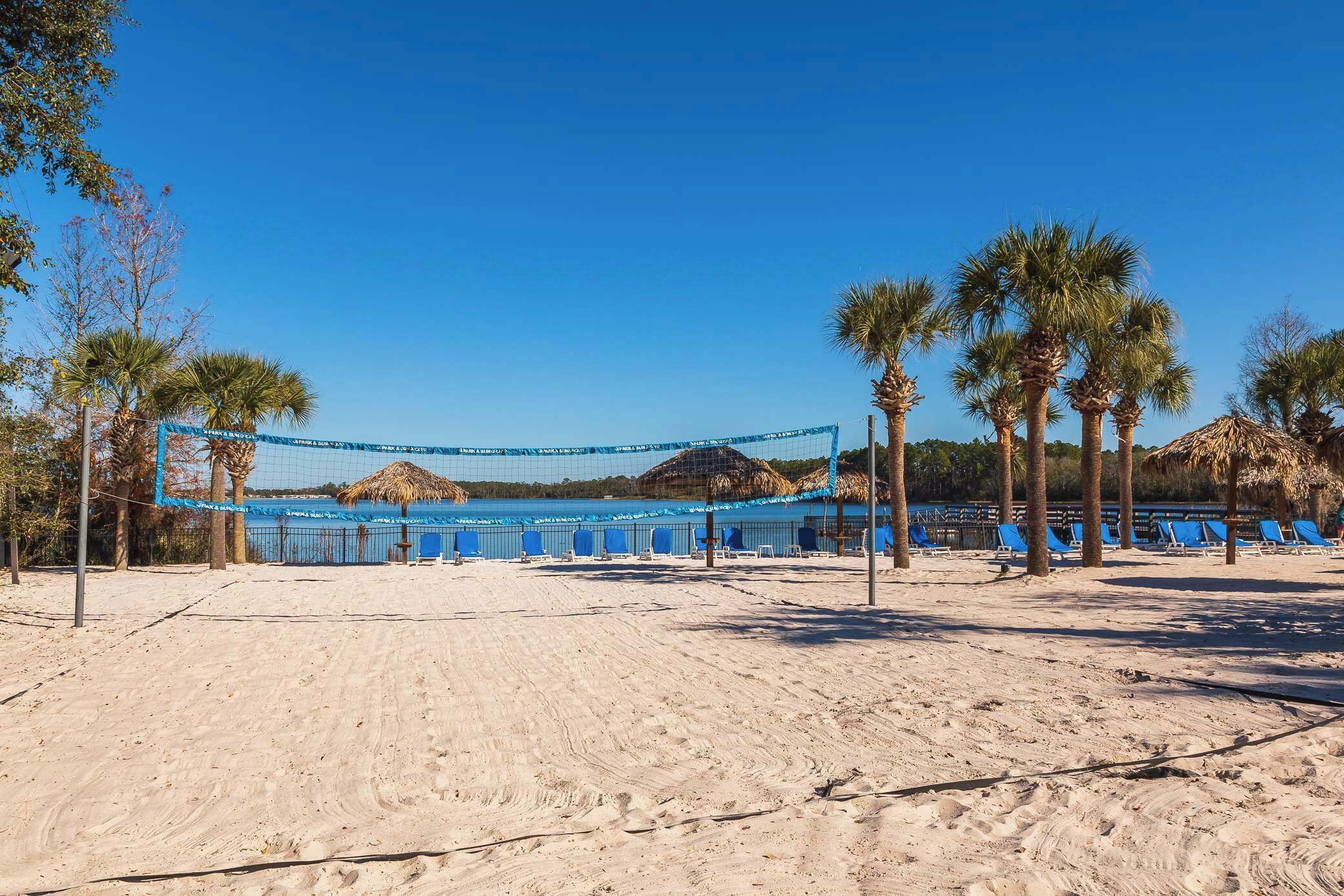 A beach volleyball court with blue netting, palm trees, and beach chairs.
