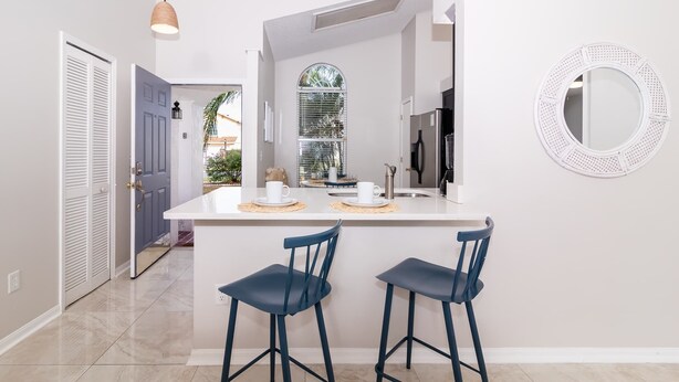 Una cocina moderna con una isla blanca, dos taburetes azules, un espejo redondo y una ventana con persianas.