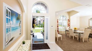 A hotel lobby with a large painting of a beach scene, a dining area with a table and chairs, and a staircase leading to a balcony. - Beachy Retreat (Kissimmee)