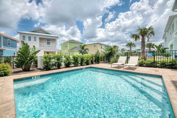 A swimming pool with a clear blue water, surrounded by a fence, and adjacent to a row of colorful houses.