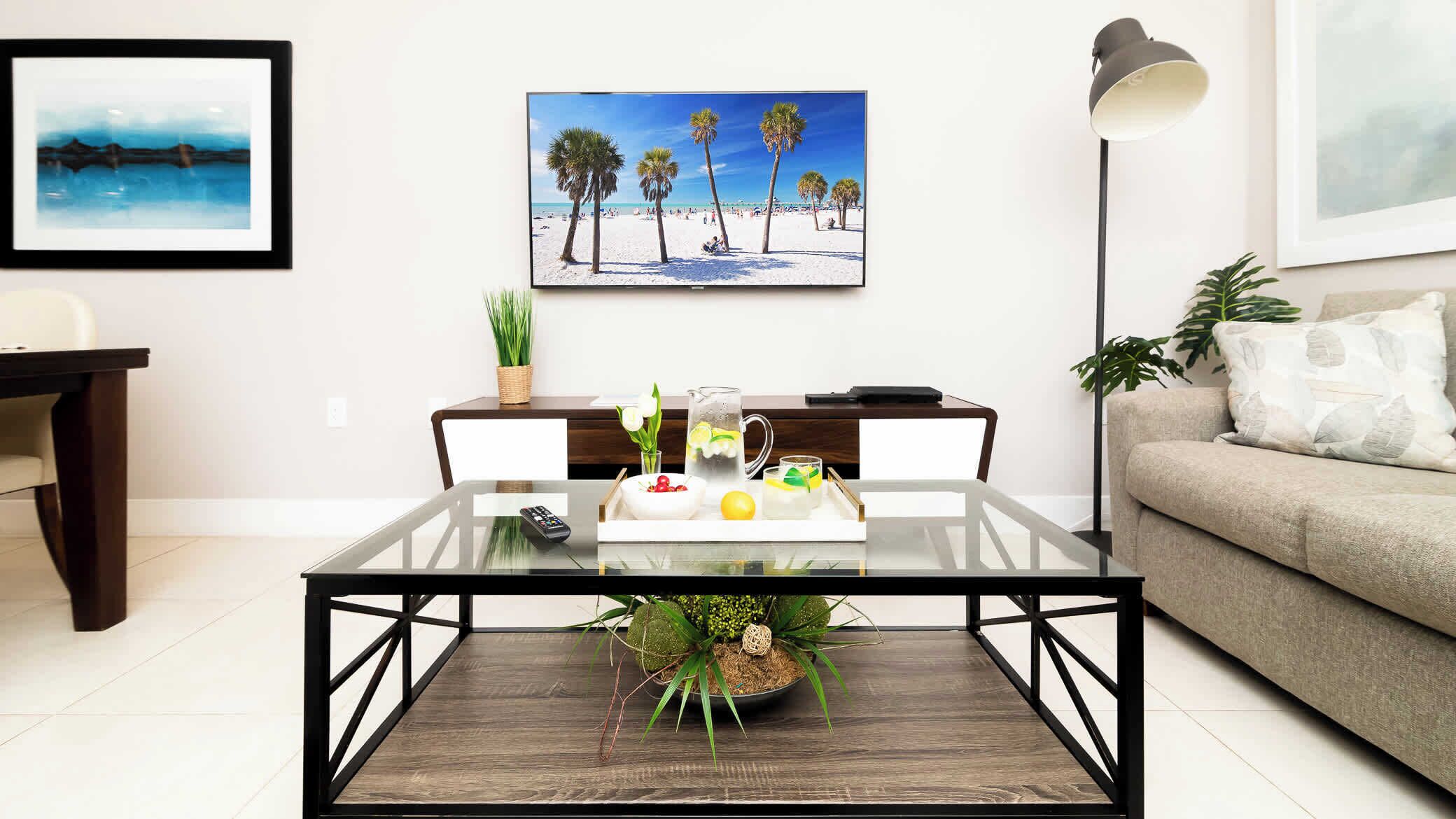 Sala de estar com mesa de centro de vidro, controle remoto, jarra de água e uma tigela de frutas. Há uma pintura de uma cena de praia na parede, um abajur e um sofá com um almofado.