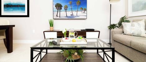 Sala de estar com mesa de centro de vidro, controle remoto, jarra de água e uma tigela de frutas. Há uma pintura de uma cena de praia na parede, um abajur e um sofá com um almofado.