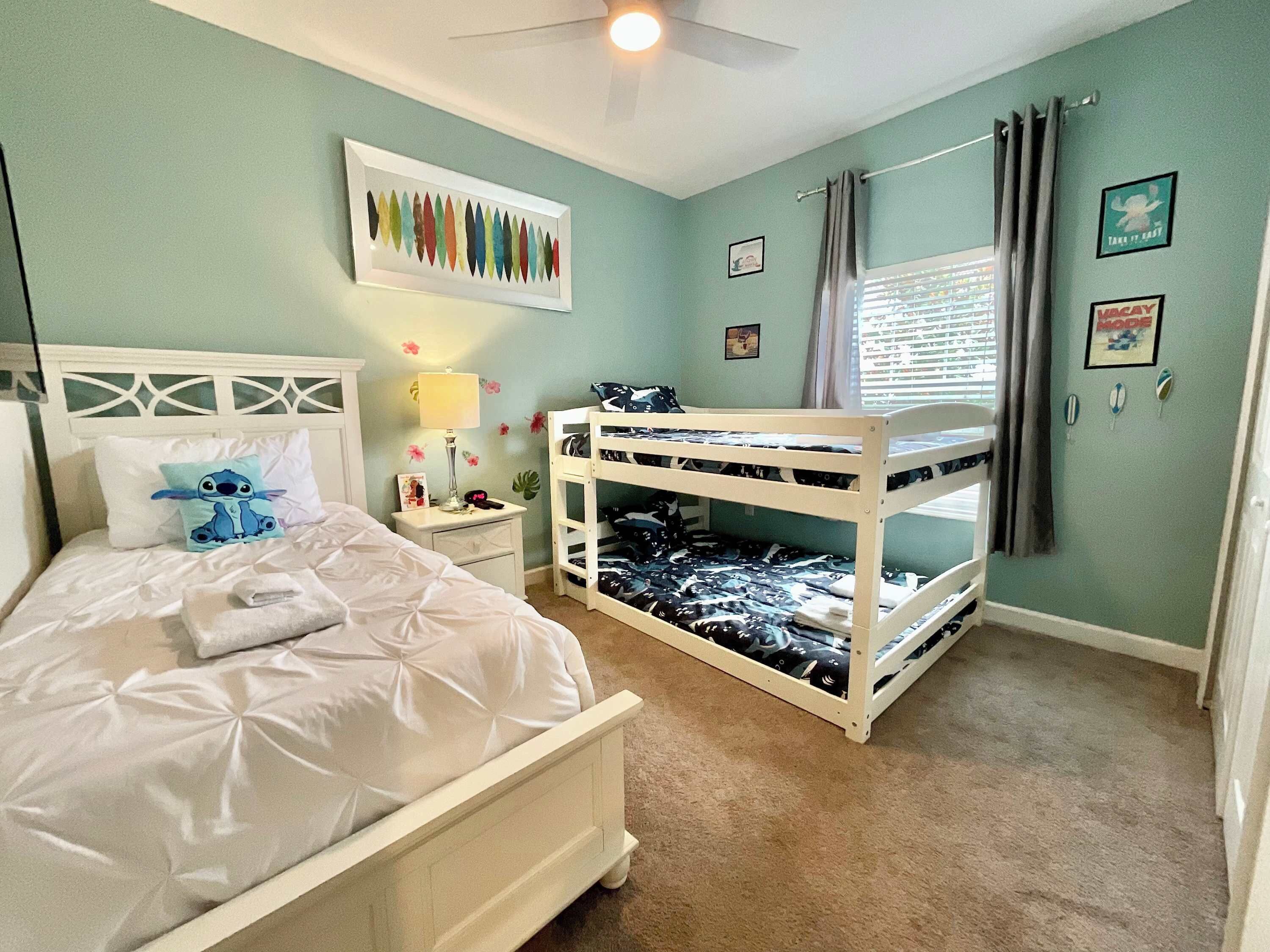 A bedroom with a bunk bed, a single bed with a pillow featuring a character, a nightstand with a lamp, and a wall with framed pictures.