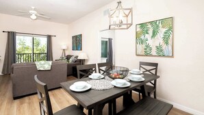 A dining area with a wooden table set for four, surrounded by dark chairs. A chandelier hangs above the table, and a large tropical leaf painting is on the wall. - Family Ties at Bahama Bay - Luxe 3BR Townhouse (Davenport)