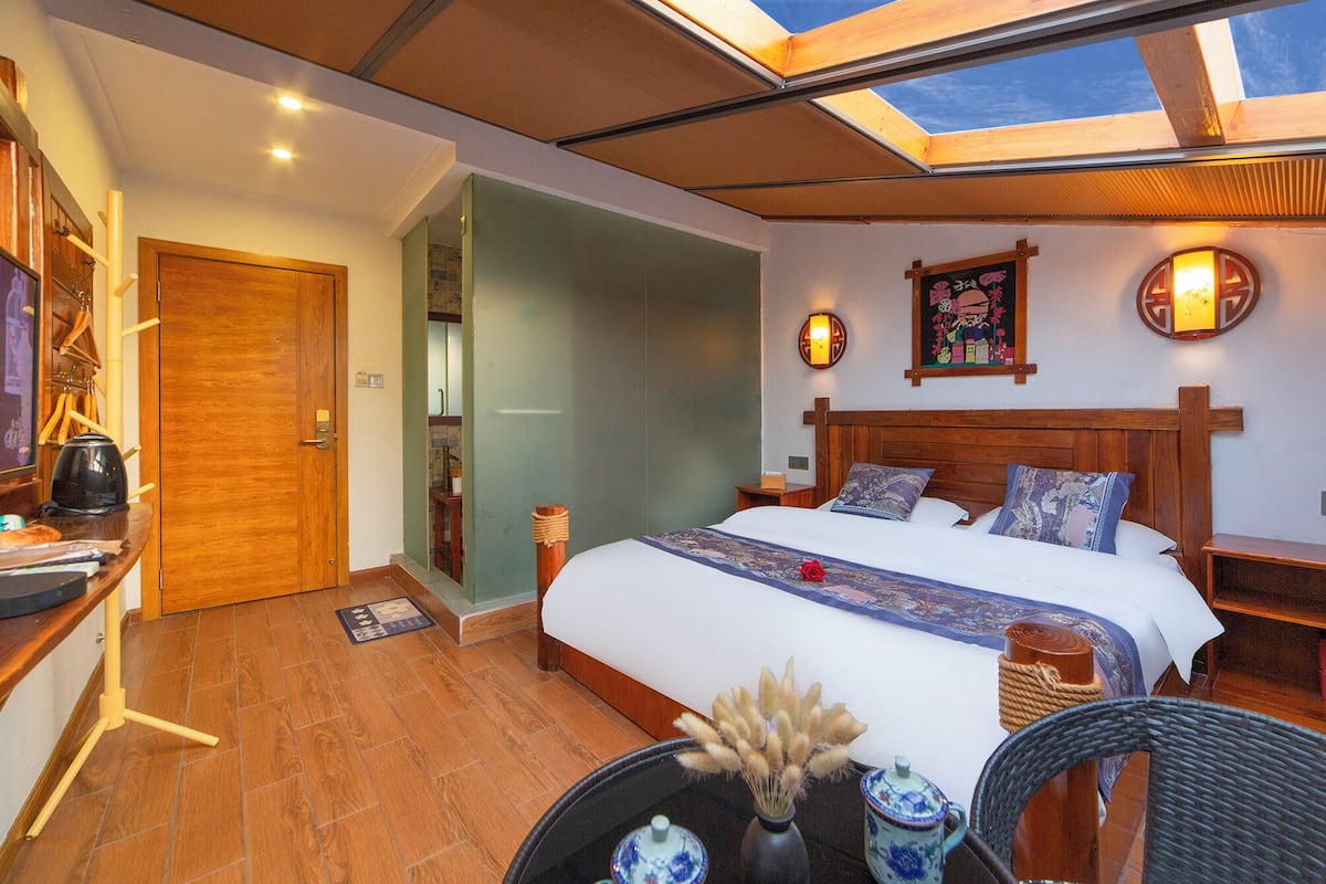 Une chambre d’hôtel avec un grand lit, une tête de lit en bois, une table de chevet, une petite table d’appoint, une lampe fixée au mur, une décoration murale et un puits de lumière.