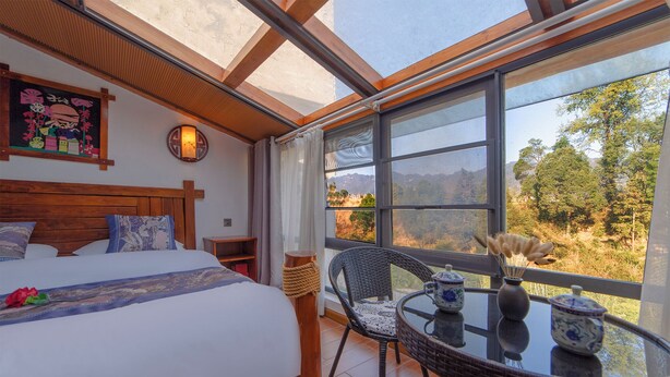 A bedroom with a large bed, a wooden headboard, a nightstand, a wicker chair, a small table with decorative items, and a view of a mountainous landscape through large windows. - Zhangjiajie Xinghe Qingmeng Folk-style Inn (Zhangjiajie)
