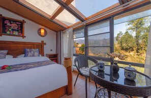 Une chambre dotée d’un grand lit, d’une tête de lit en bois, d’une table de chevet, d’un fauteuil en osier, d’une petite table ornée d’objets décoratifs et offrant une vue sur un paysage montagneux grâce à de grandes fenêtres.