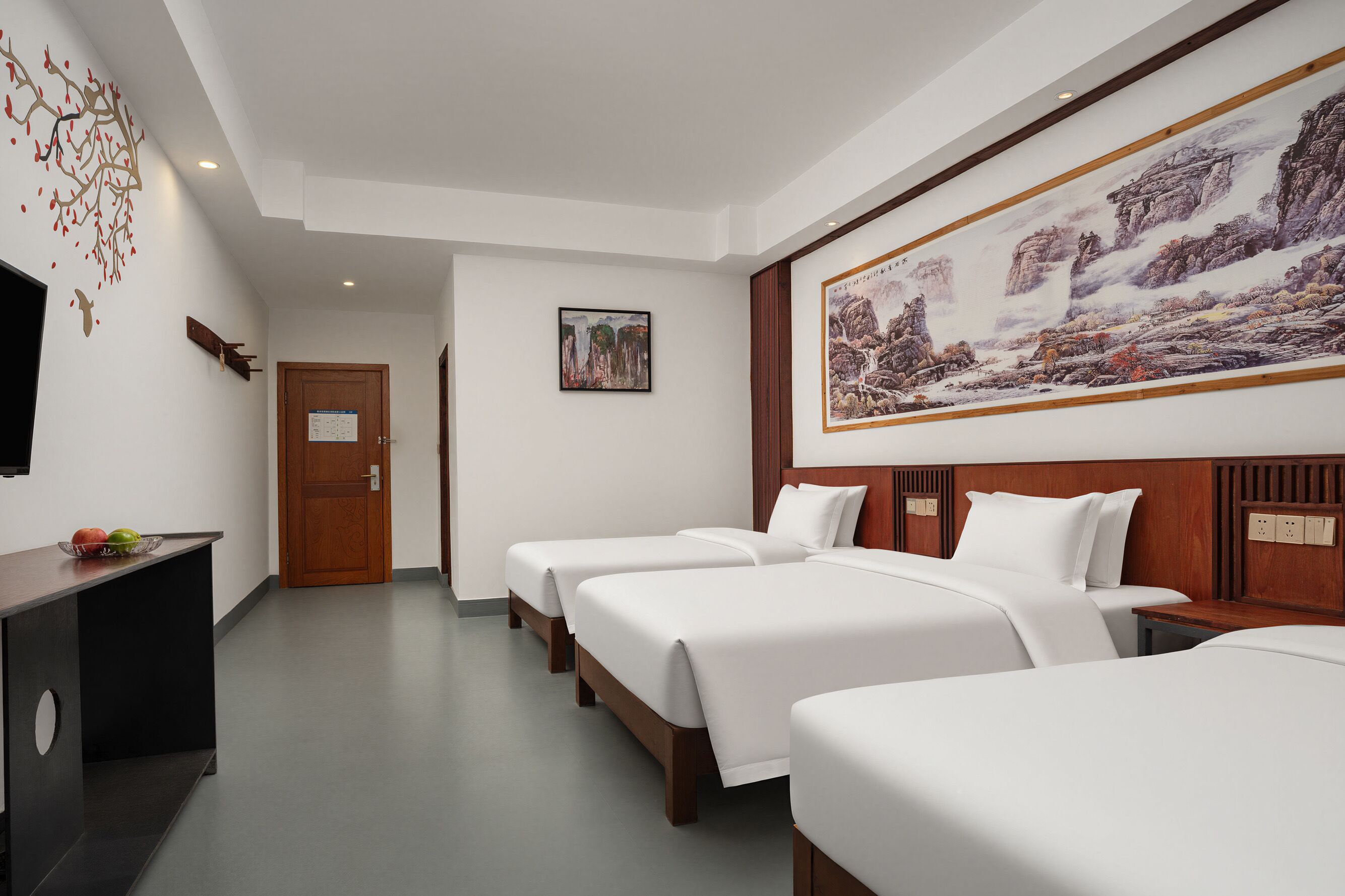 A hotel room with three beds, a wooden headboard, a flat-screen TV, a wall-mounted artwork, and a wooden desk with a fruit bowl.