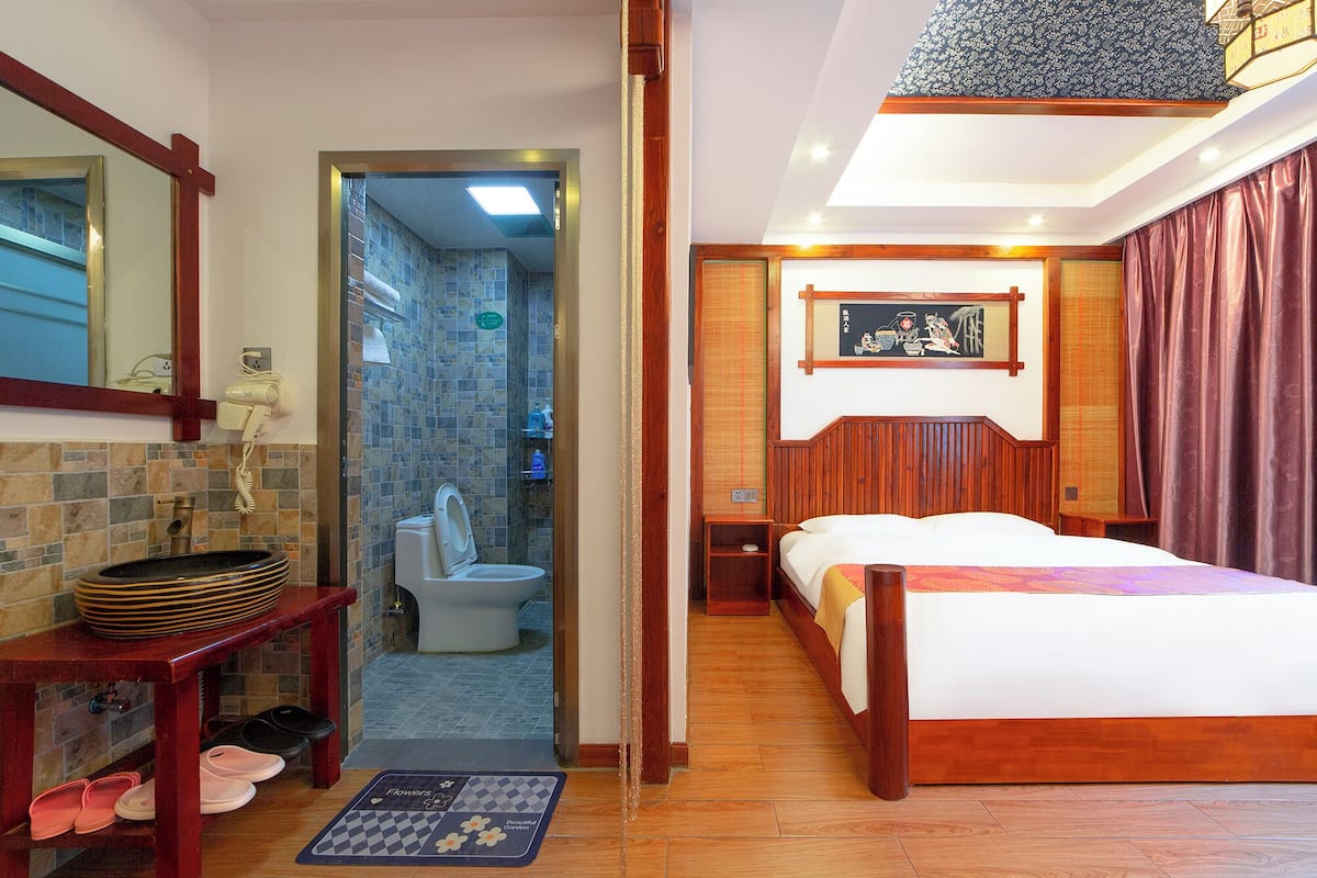 Une chambre d’hôtel avec un lit en bois, une salle de bain avec douche et une petite table avec des chaussons.
