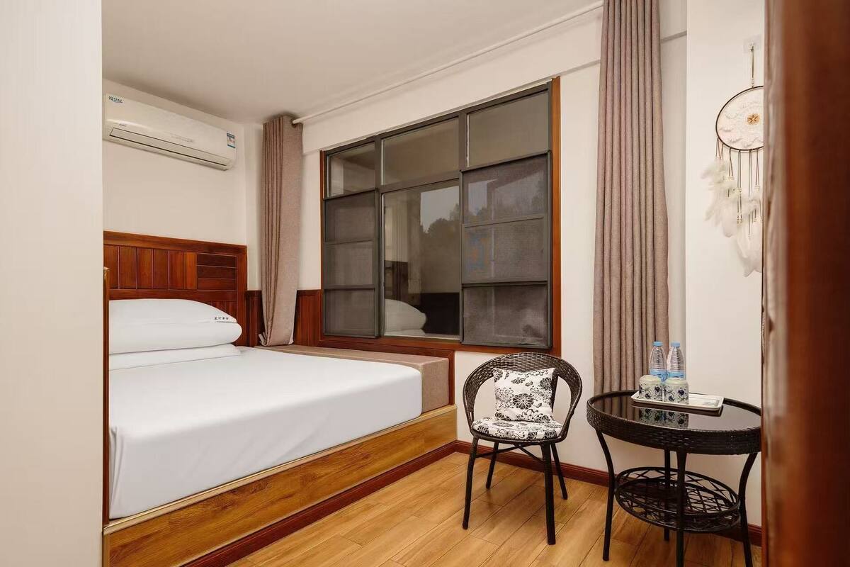 Une chambre d’hôtel avec un lit, une fenêtre, une chaise, une petite table avec des bouteilles et un attrape-rêves fixé au mur.