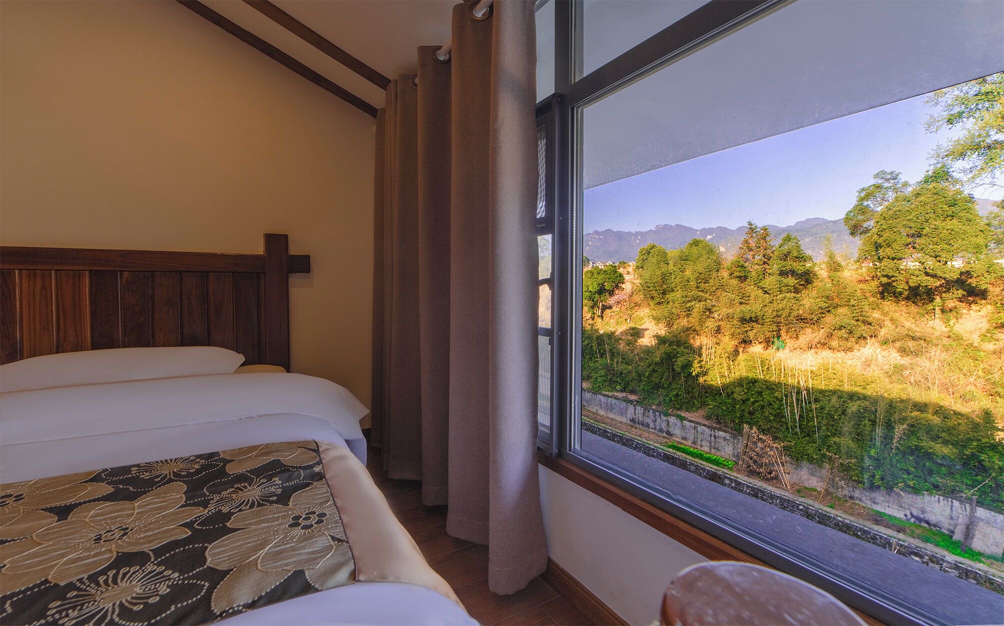 A hotel room with a bed, a window with a view of a forest and mountains, and a wooden headboard.