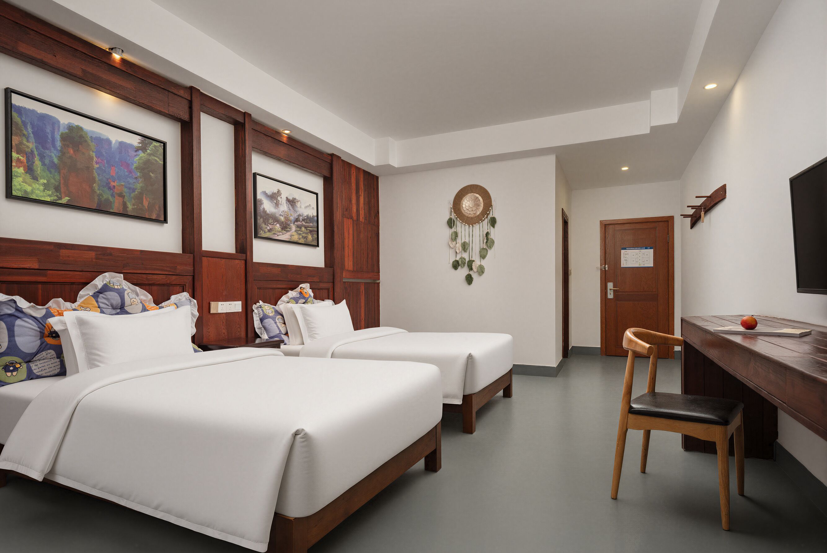 A hotel room with two beds, a desk, a chair, and a dreamcatcher on the wall.