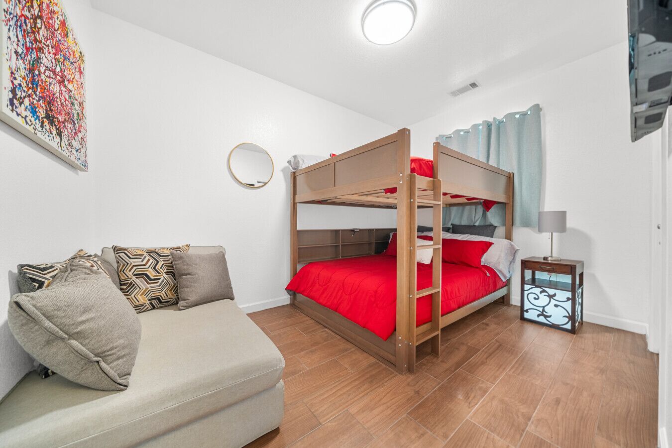 A bunk bed with red bedding, a sofa with patterned cushions, a small bedside table with a lamp, and a colorful abstract painting on the wall.