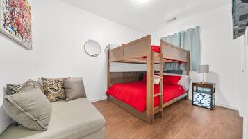 A bunk bed with red bedding, a sofa with patterned cushions, a small bedside table with a lamp, and a colorful abstract painting on the wall.