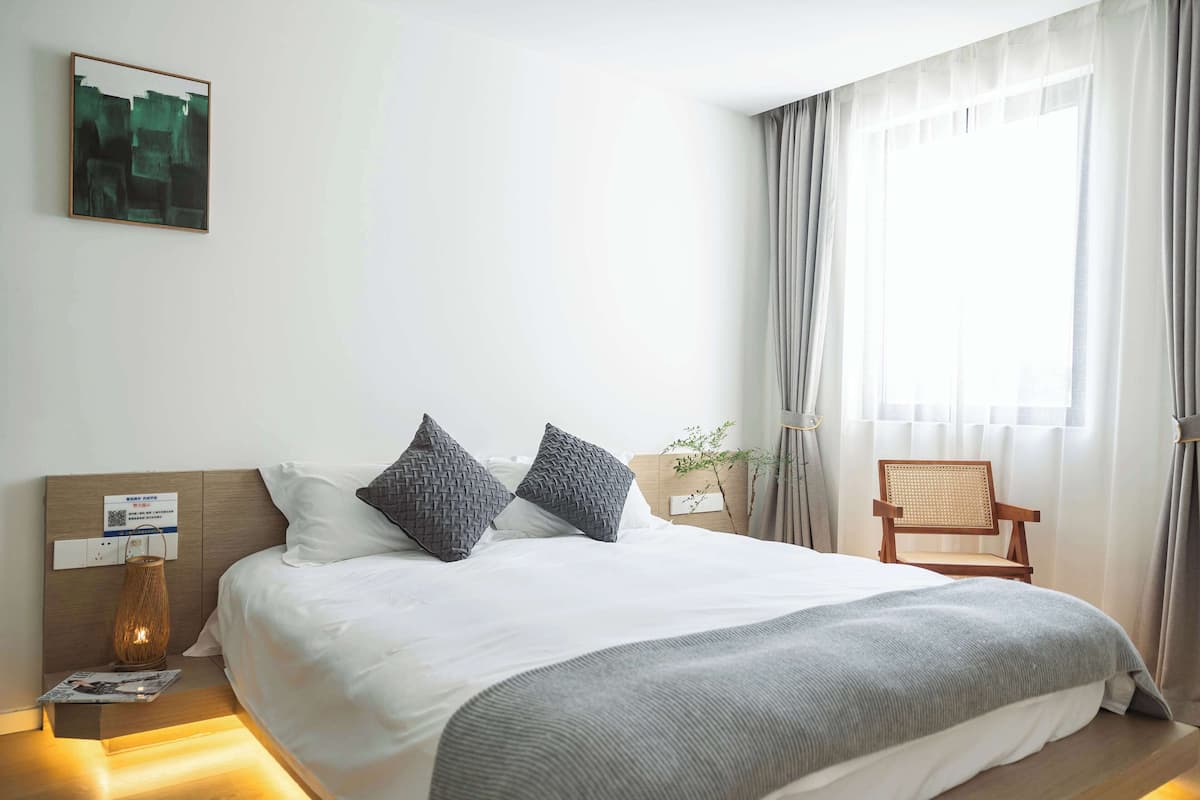 A modern hotel room with a neatly made bed, a bedside table with a lamp, a chair, and a window with curtains.