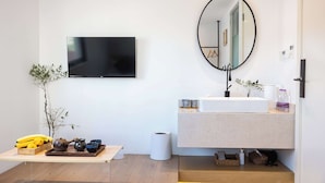 Un baño moderno con una televisión de pantalla plana, un espejo redondo, un lavabo y una estantería de madera con una planta y una bandeja de plátanos.