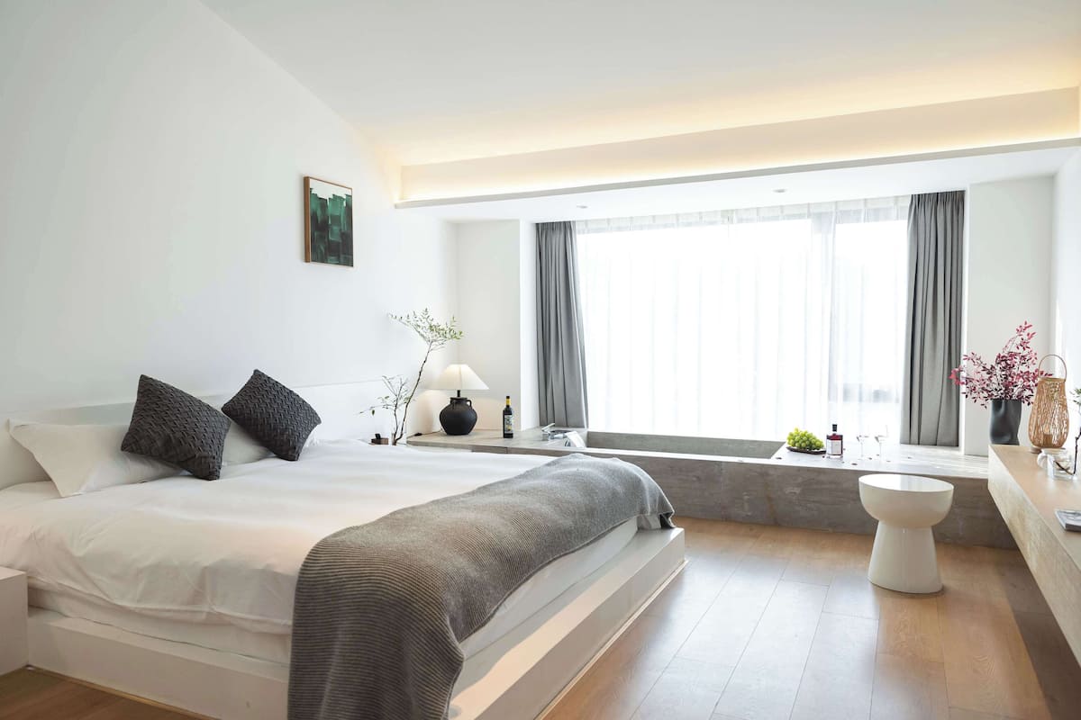 A modern bedroom with a large bed, a bedside table, a lamp, a window with curtains, and a bathroom with a toilet and sink.