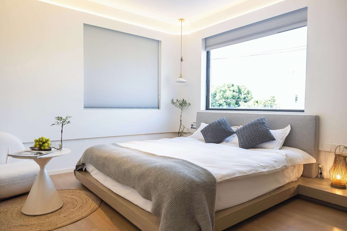A modern bedroom with a large bed, bedside tables, a small table with a plant, and a window with a view of greenery.