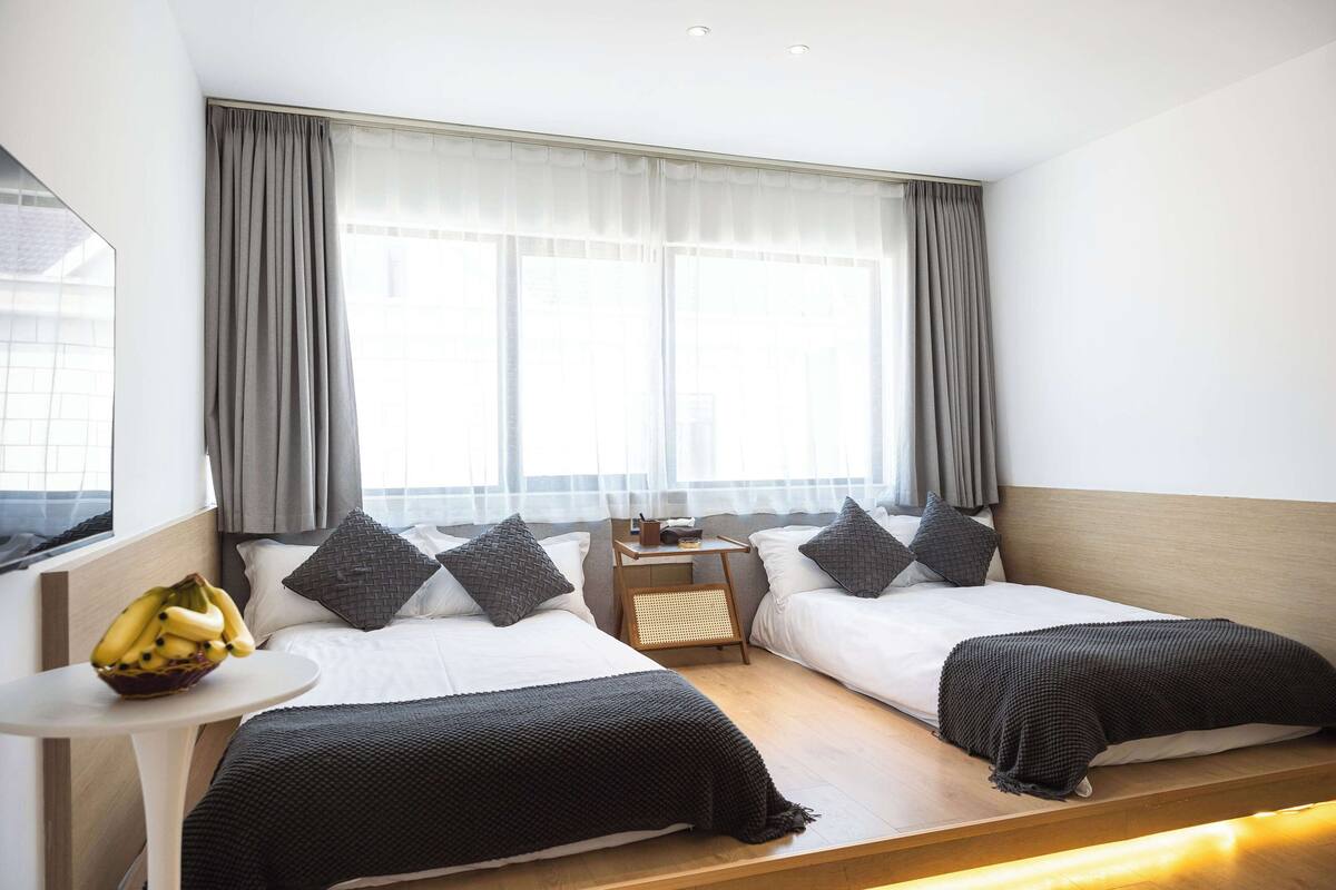 A hotel room with two beds, a small table with a fruit bowl, a nightstand, and a window with curtains.