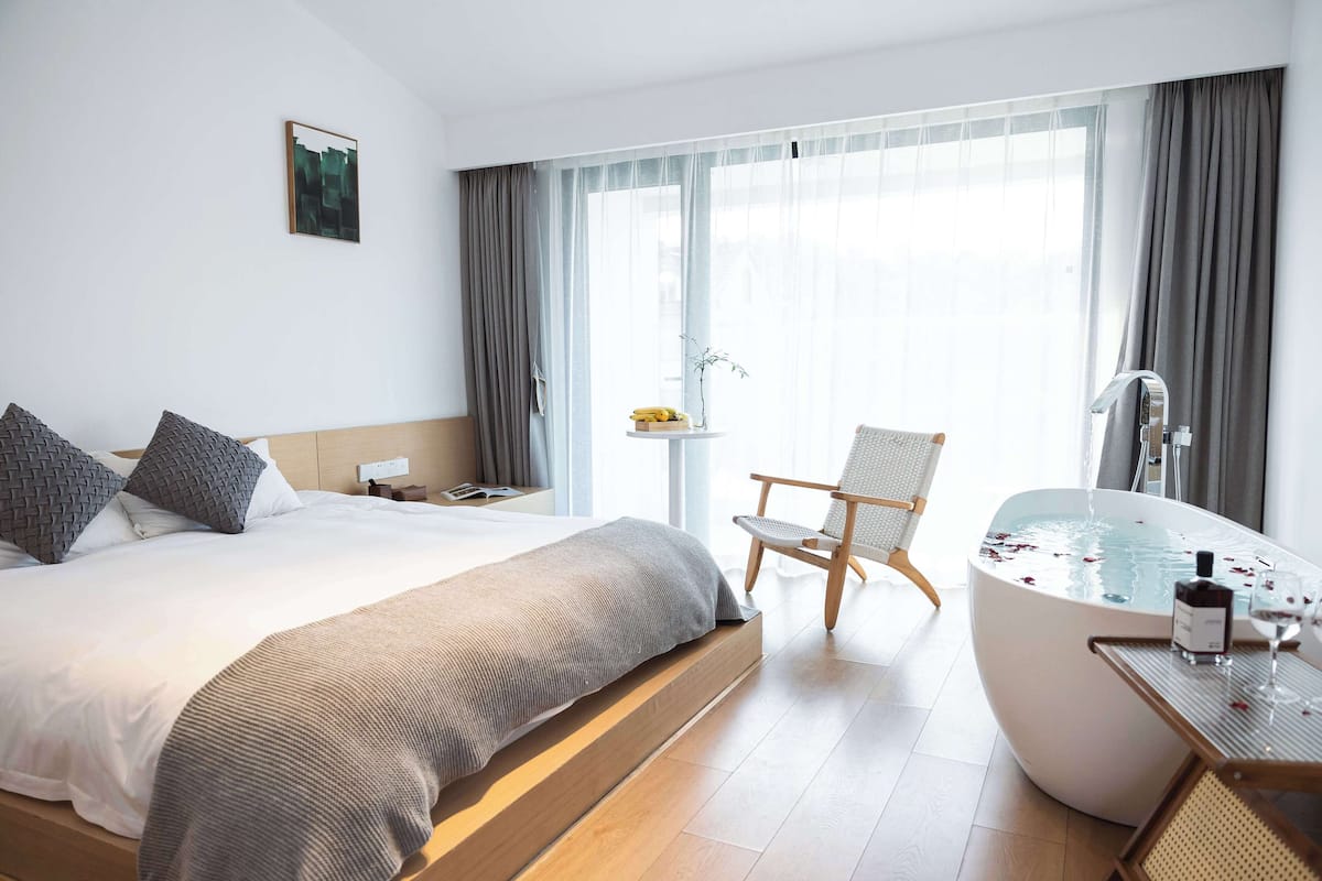 A modern hotel room with a large bed, a sitting area with a chair, a bathtub, and a small table with a bottle and glass.