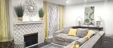 A modern living room with a sectional sofa, a fireplace, decorative plants, and a large abstract painting.
