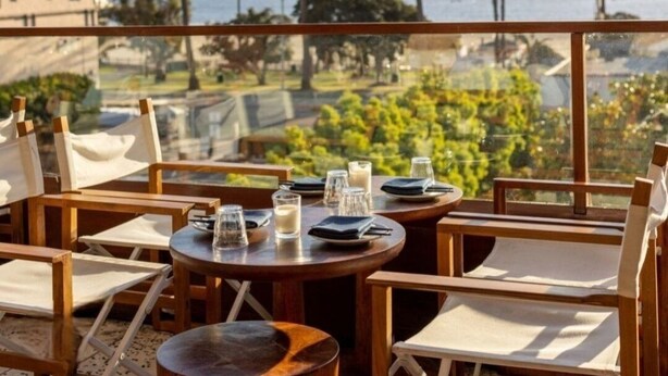 A terrace with wooden tables and chairs, overlooking a coastal landscape with palm trees and the sea. - Beach • Pools • Comfort:  Private Room + Bath Prime LA Location (Los Angeles)
