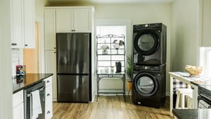 Una cocina con un refrigerador de acero inoxidable, lavadora y secadora apiladas, y una ventana con una estantería metálica.