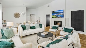Un salón moderno con un sofá blanco, cojines verdes, una chimenea y una pantalla grande de televisión mostrando un paisaje.