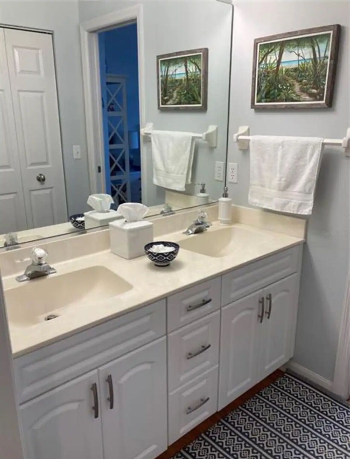 A bathroom with a double sink, white cabinets, a mirror, and two framed pictures on the wall.