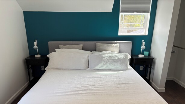 A neatly made bed with white bedding in a room with teal walls, two bedside tables with lamps, and a window with blinds. - Parkside Nest Apartment Near Manito Park (Spokane)