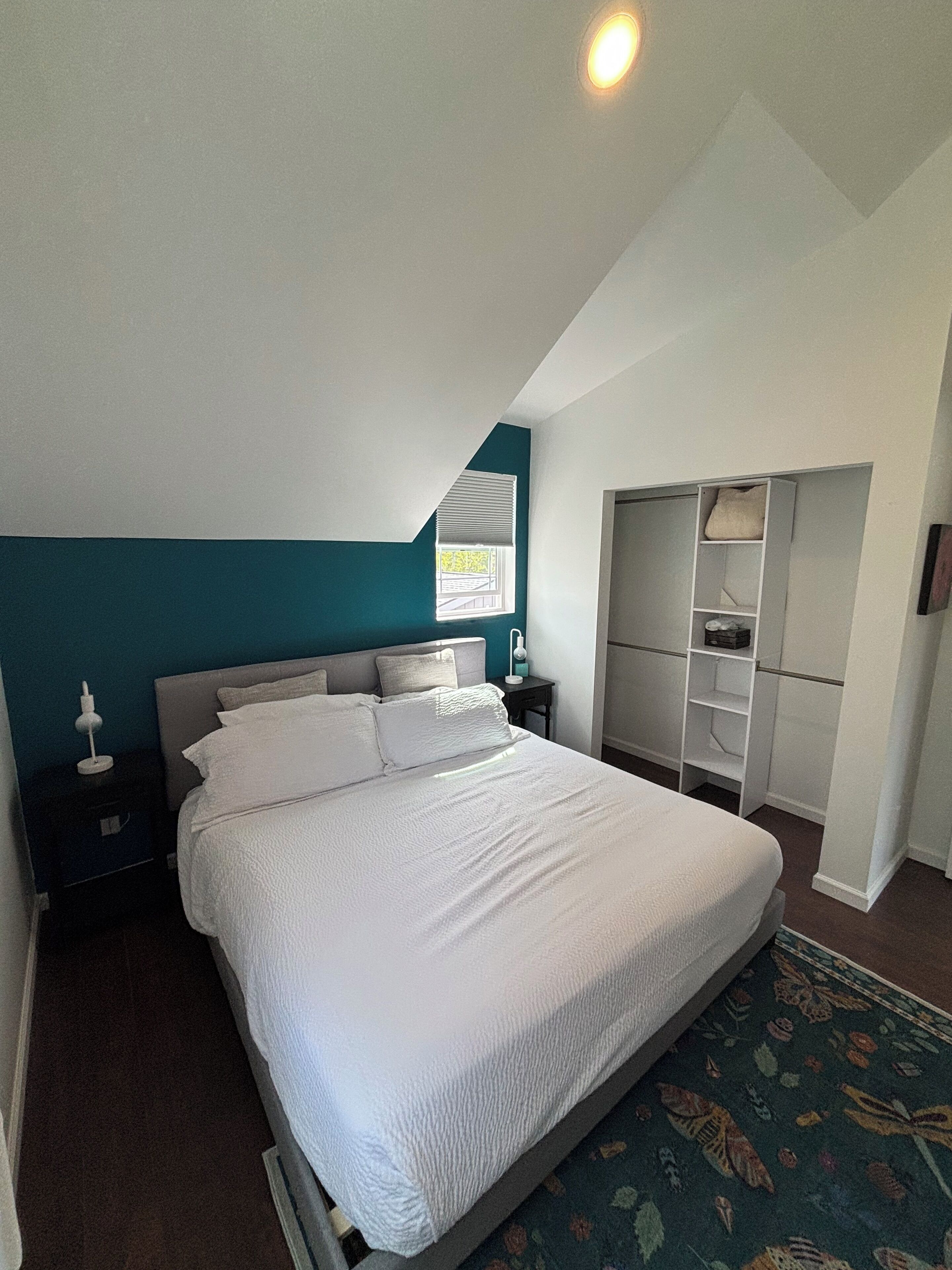 A bedroom with a large bed, a nightstand, a wardrobe, and a window with blinds.