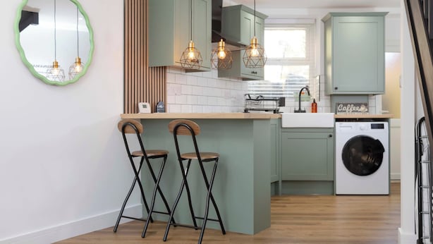A modern kitchen with green cabinets, a central island with bar stools, pendant lighting, and a built-in washing machine. - Cosy-cute Miner's Cottage on the Taff Trail (Merthyr Tydfil)