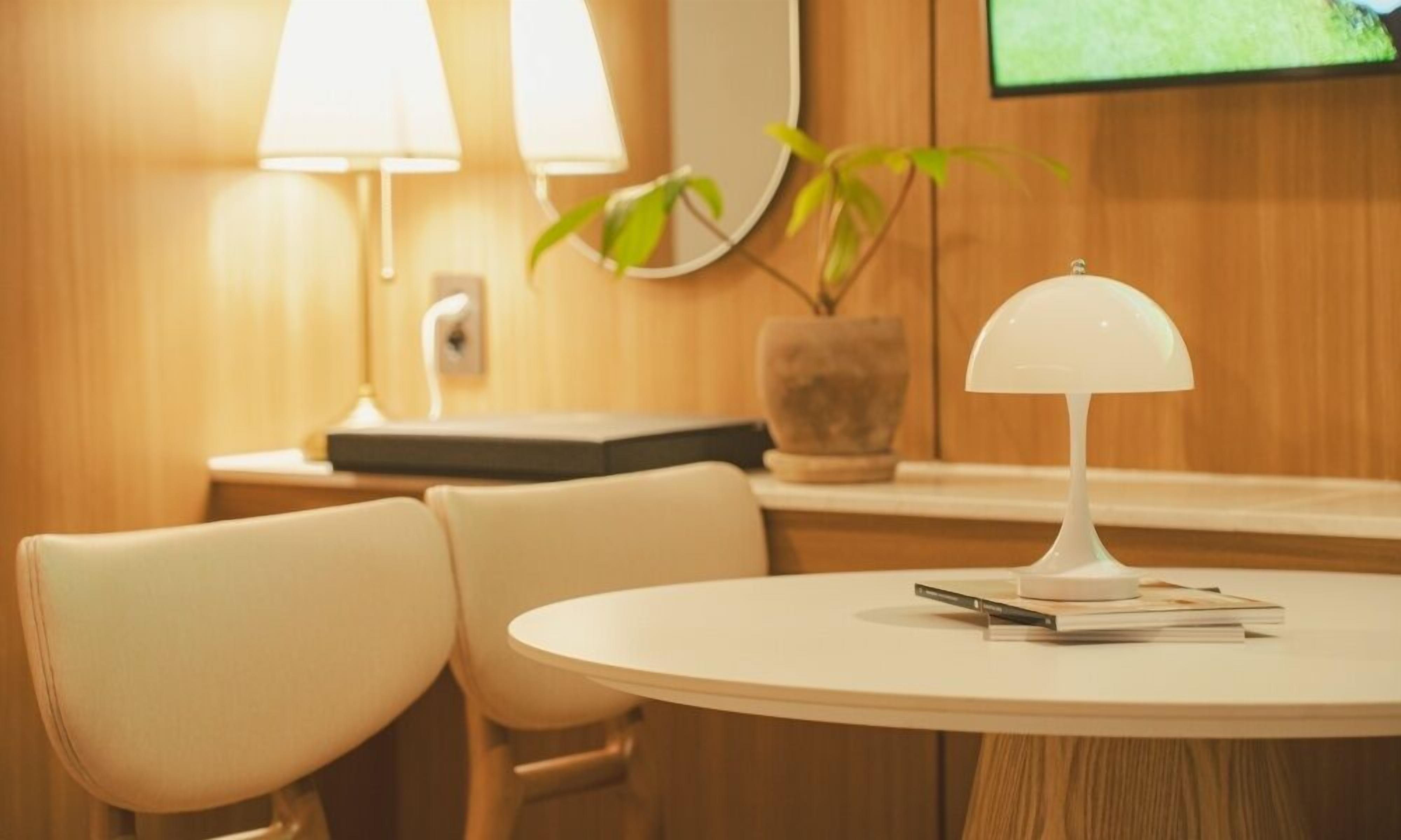 Una mesa redonda con dos sillas, una lámpara blanca, una planta y un televisor montado en la pared.