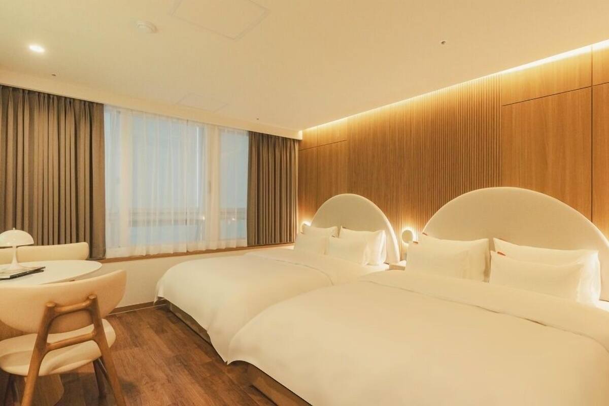 Habitación de hotel con dos camas, un escritorio con silla y un ventanal con cortinas.