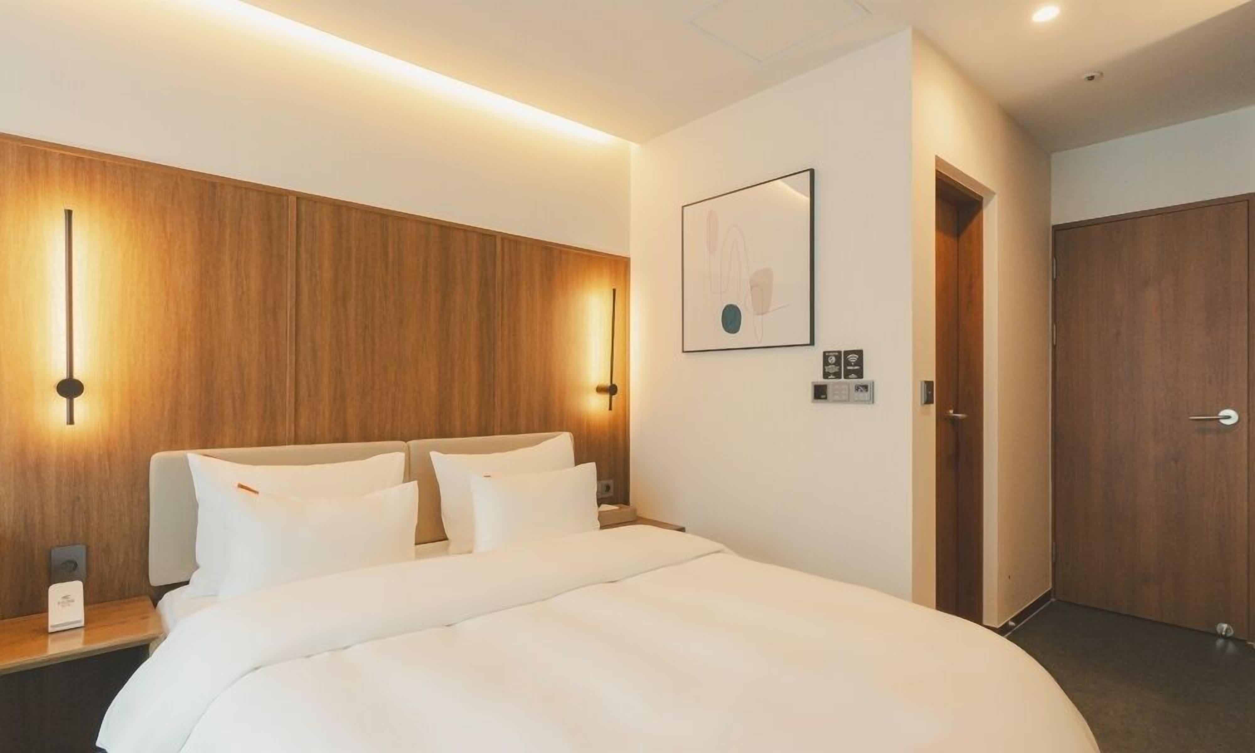 A hotel room with a large bed, wooden paneling on the walls, a wall-mounted light, and a door.