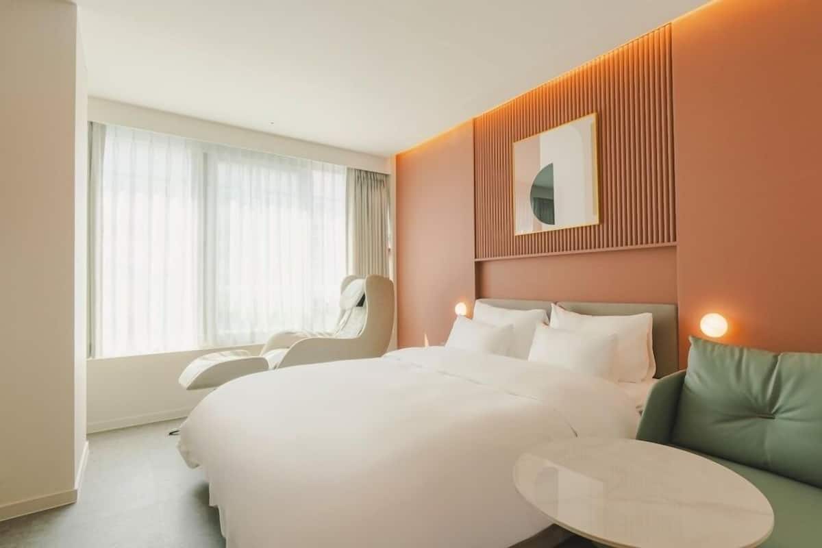 Habitación de hotel con una cama grande, una silla, una mesa redonda y vistas al exterior a través de cortinas translúcidas.