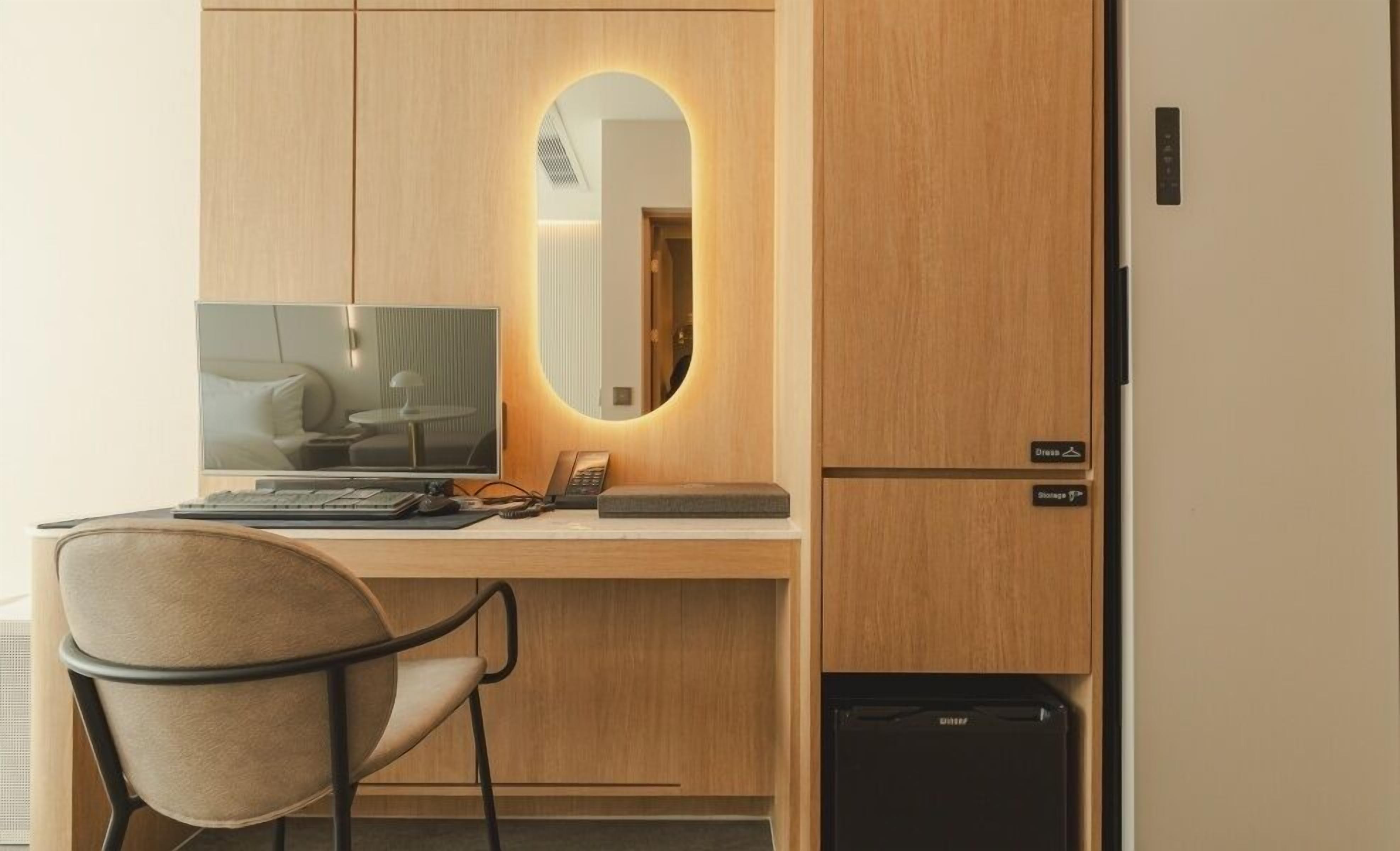 Una habitación de hotel compacta con escritorio de madera, una computadora, una silla, un espejo y un refrigerador.