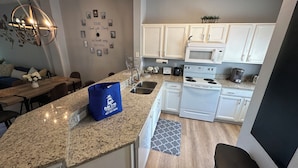 Una cocina moderna con armarios blancos, encimera de granito, horno empotrado y microondas. Hay una bolsa azul sobre la encimera y un reloj en la pared.