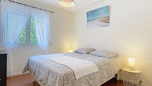 Een slaapkamer met een bed, een bedlamp, een raam met dunne gordijnen, een hanglamp en een schilderij aan de muur.
