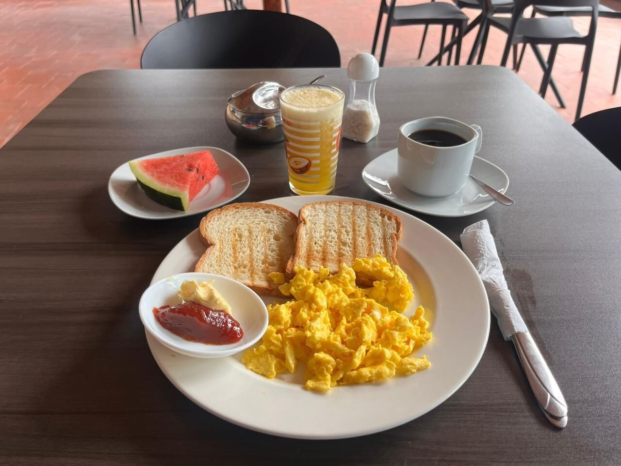 A breakfast plate with scrambled eggs, toast, a slice of watermelon, a glass of juice, and a cup of coffee on a wooden table.
