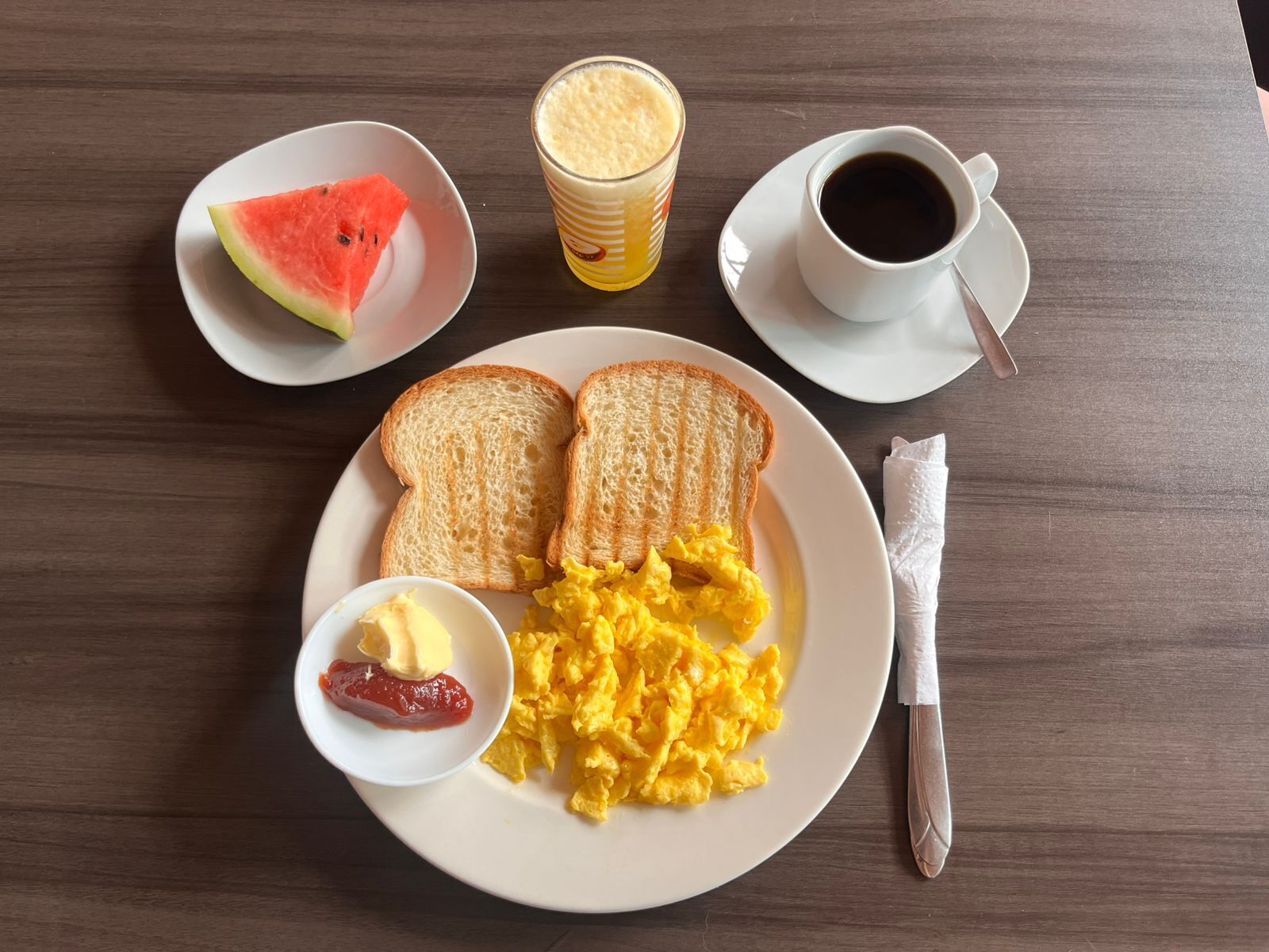 A breakfast plate with scrambled eggs, toast, butter, jam, a glass of orange juice, and a cup of coffee on a wooden table.