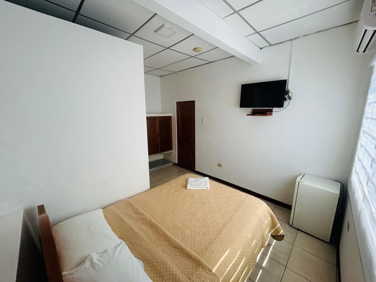 A hotel room with a bed, a television mounted on the wall, and a small air conditioning unit.