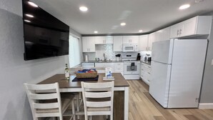 A modern kitchen with white cabinets, a built-in microwave, a refrigerator, and a dining area with two chairs and a table. - Piping Plover on the Key, Nautical and Nice, Paddles away! (Englewood)